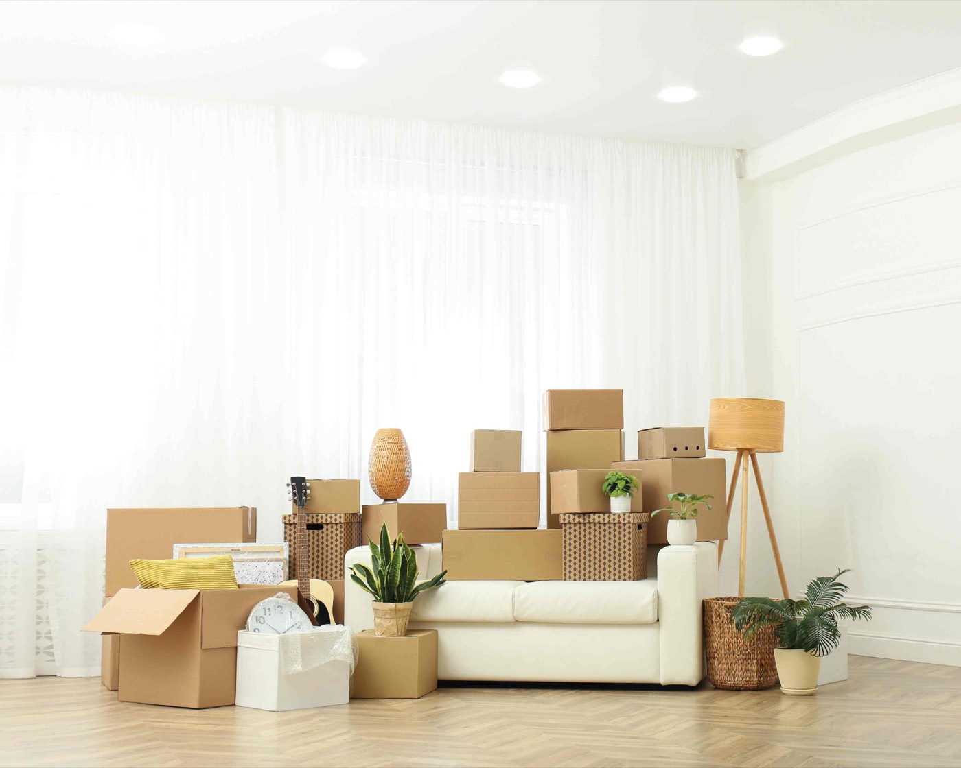 Packed living room ready for a move, with a sofa surrounded by stacked moving boxes, a guitar and houseplants.